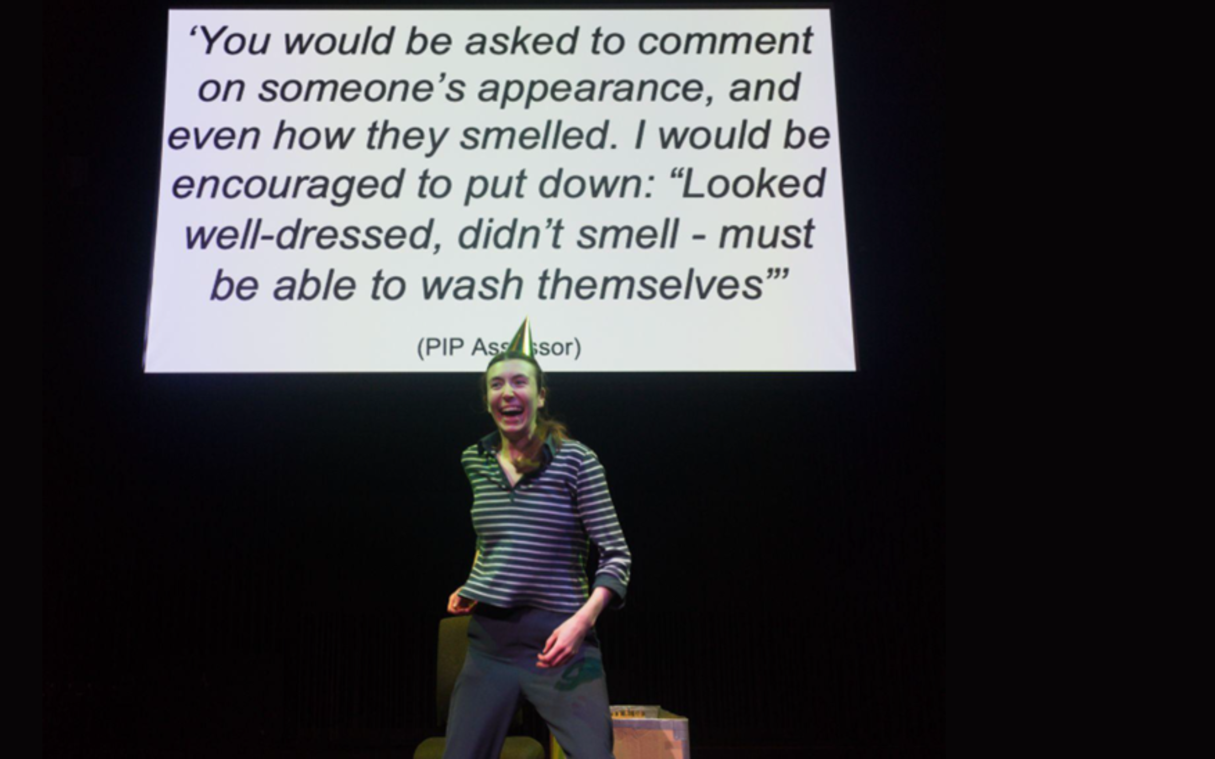 A performer wears a stripey black and white top with a gold party hat. She is smiling broadly. There is a white screen behind her which shows a quote in black text.