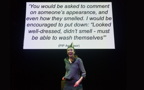 A performer wears a stripey black and white top with a gold party hat. She is smiling broadly. There is a white screen behind her which shows a quote in black text.