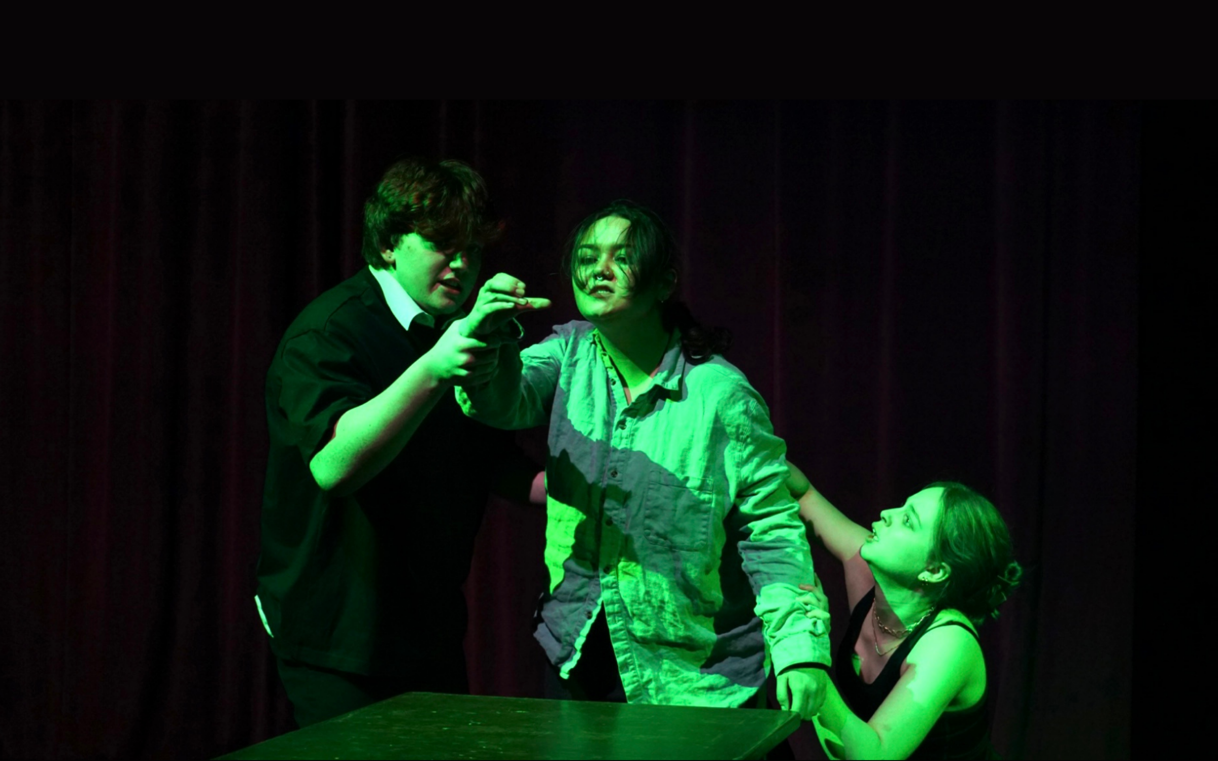 Three performers stand behind a table. They are lit in an eerie green light and the central performer prepares to take a drink.