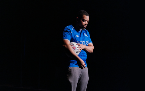A photo of a young man holding a football and wearing football kit on a black stage.