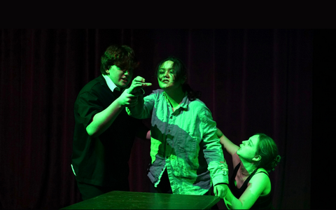 Three performers stand behind a table. They are lit in an eerie green light and the central performer prepares to take a drink.