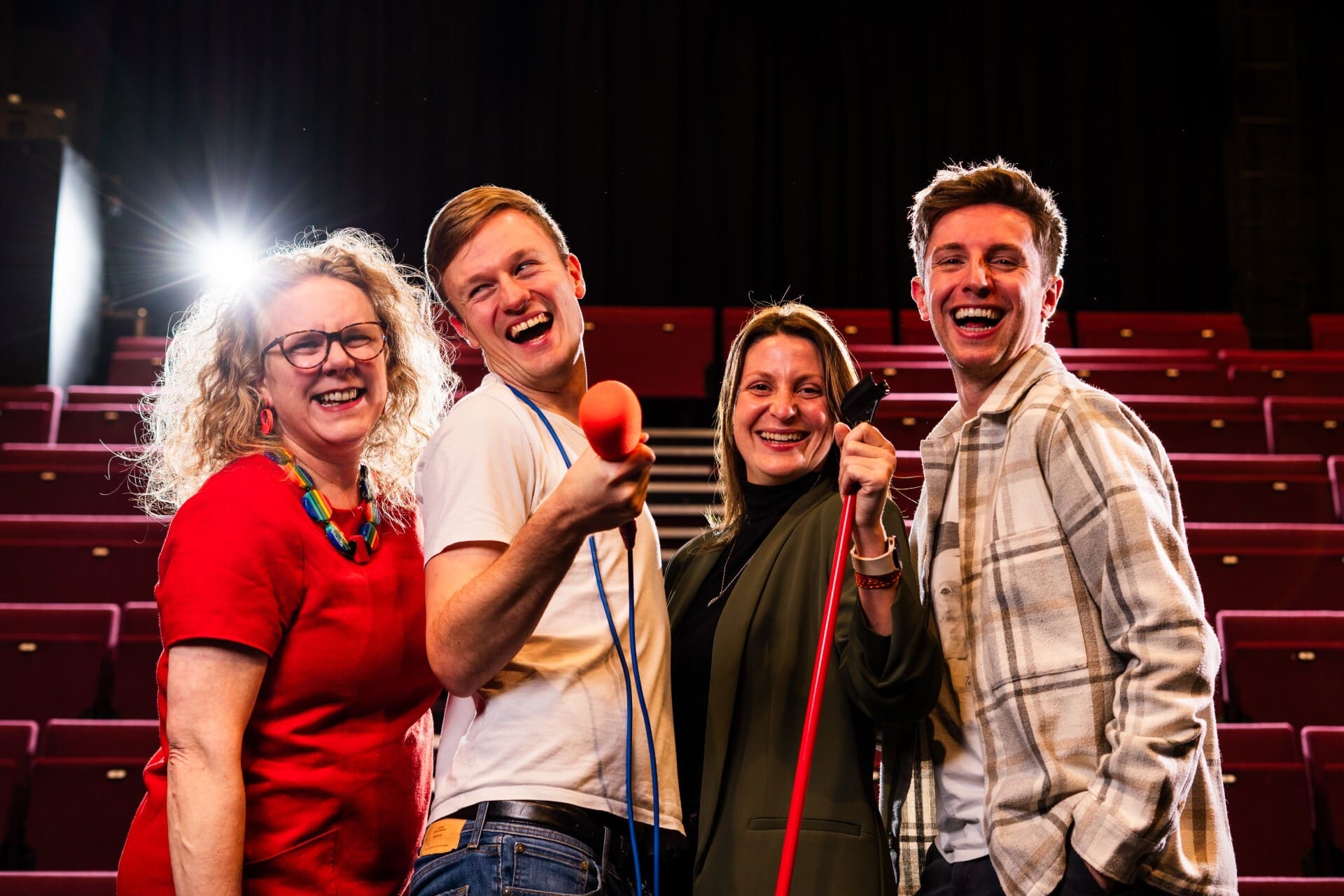 Four people stand in an empty theatre holding a microphone and a red cane, facing the camera with stage lights behind them.