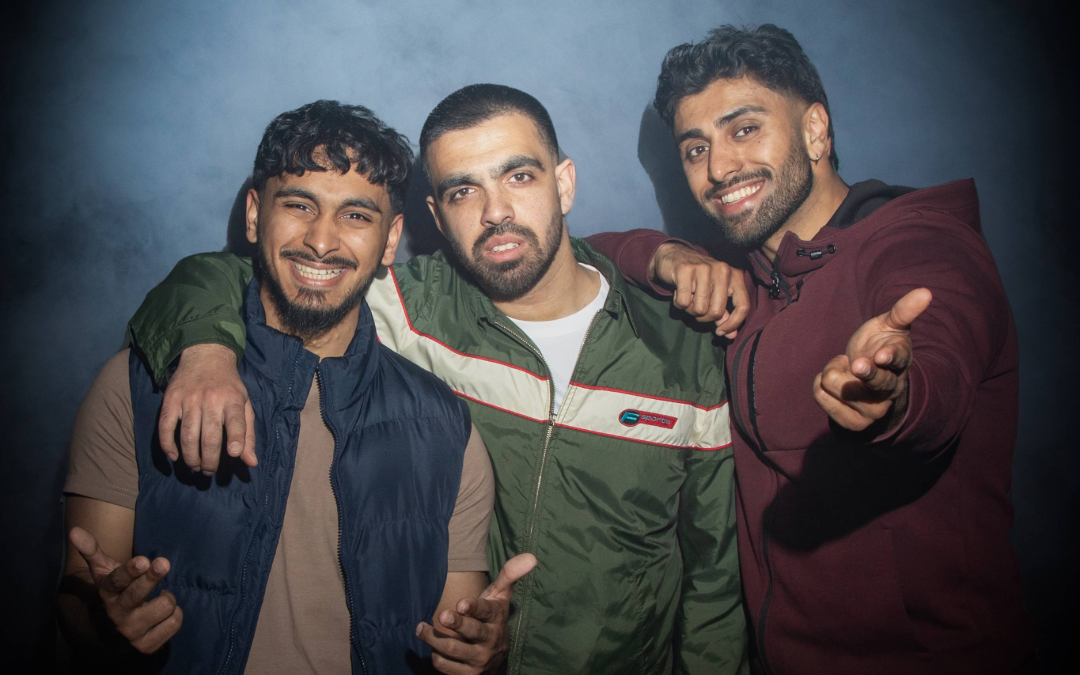 Three men standing close together in casual jackets with a smoky, dark background, posing with expressive hand gestures.