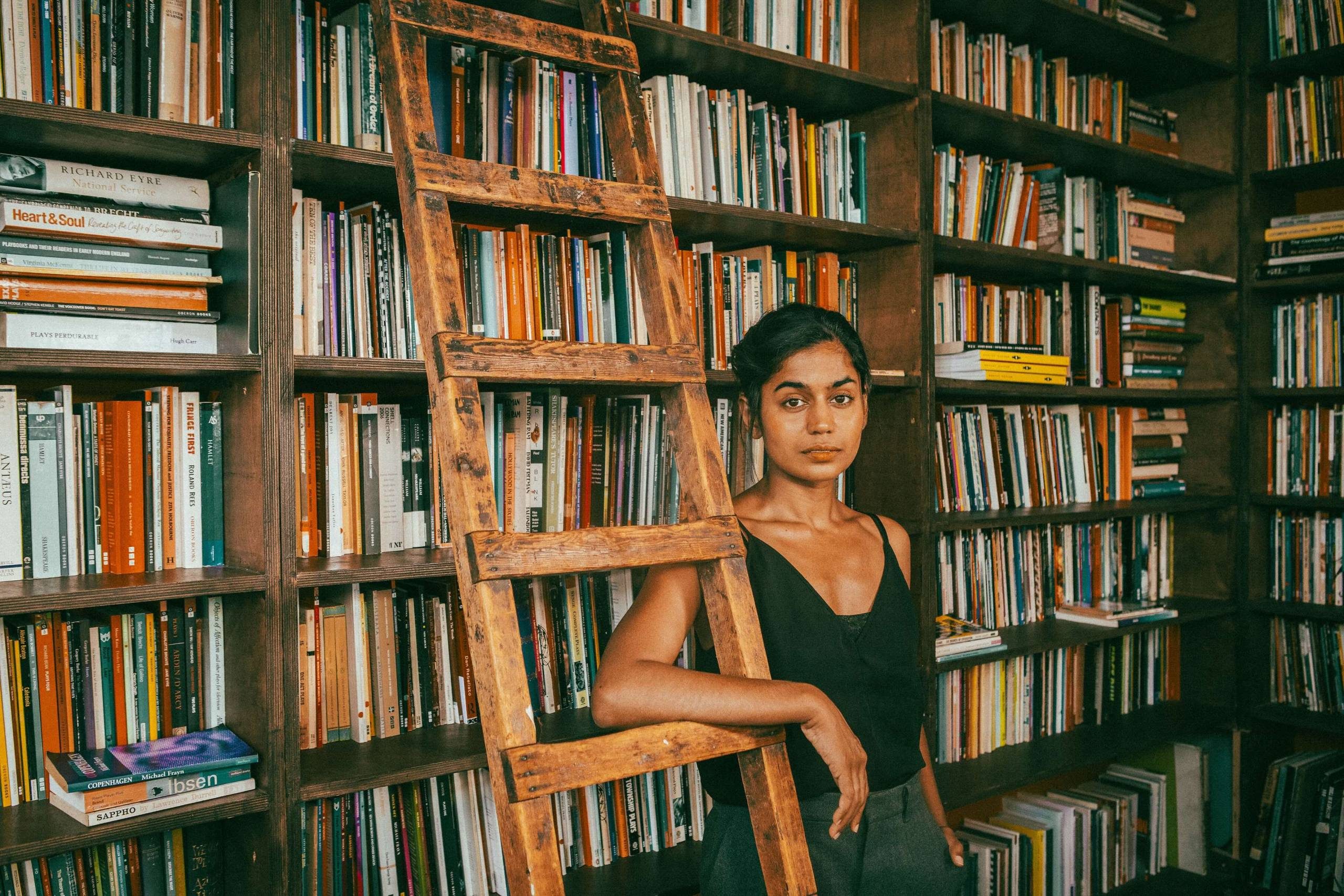 Linnea Berthelsen stands beside a wooden ladder in a library filled with numerous bookshelves. The shelves are packed with a variety of books. The atmosphere is calm and scholarly.