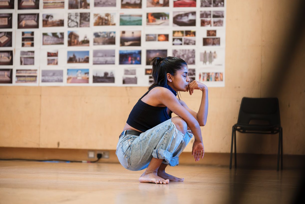 Linnea Berthelsen is thoughtfully squatting in a room with many photographs on the wall. She is barefoot, wearing a black top and blue jeans. An empty chair is visible nearby.