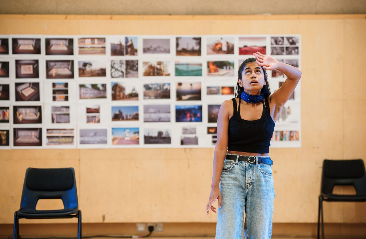 Linnea Berthelsen stands looking upward thoughtfully, with one hand on her forehead. She is wearing a dark tank top and light blue jeans. Behind them, a wall displays a collection of photographs arranged in a grid. Two empty chairs are positioned on either side of her.