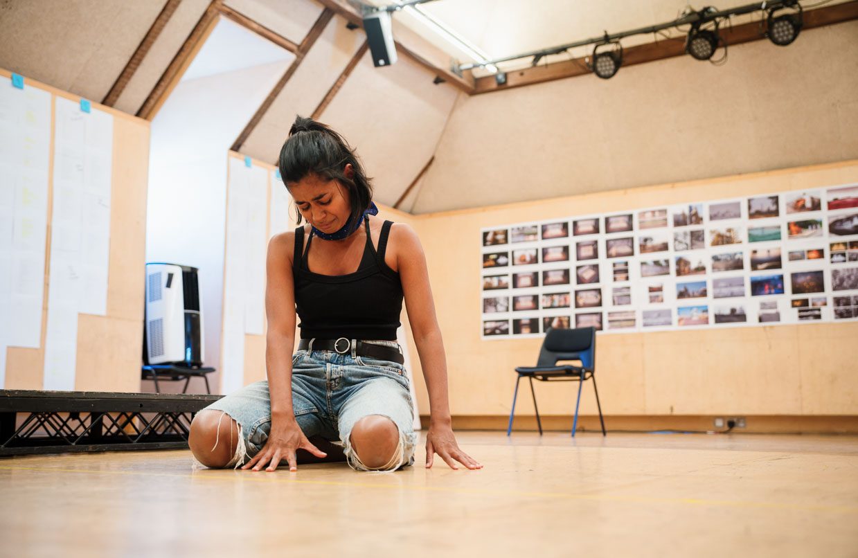 Linnea Berthelsen crying as she kneels on the floor of the rehearsal room. The room has design sketches posted on one wall and a chair nearby. She is wearing a black top and blue ripped jeans.
