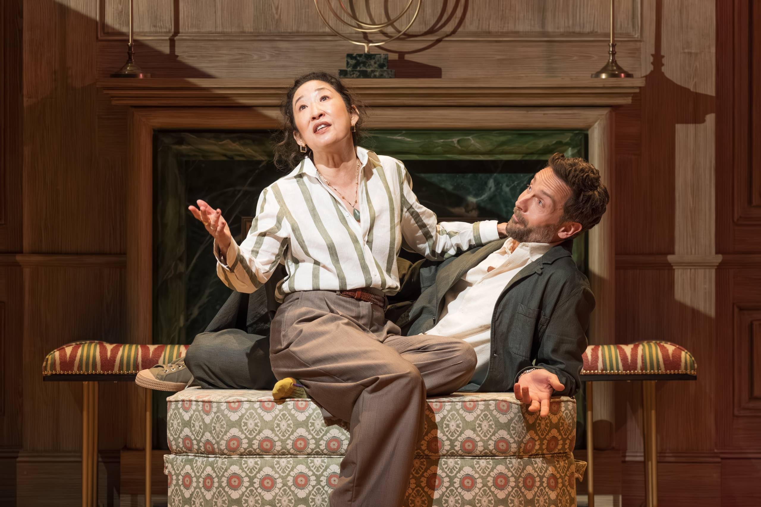 Two people are in a theatrical scene on stage, with one person sitting on an ottoman and another reclining beside them. They are in a warmly lit room, featuring a decorative fireplace and patterned textiles.
