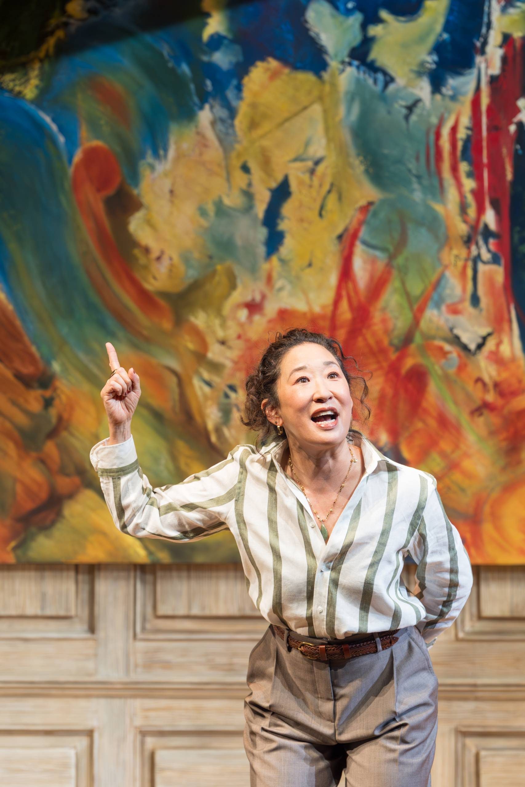 Sandra Oh standing in front of a large vibrant, abstract painting, pointing upwards, wearing a striped shirt and grey pants.