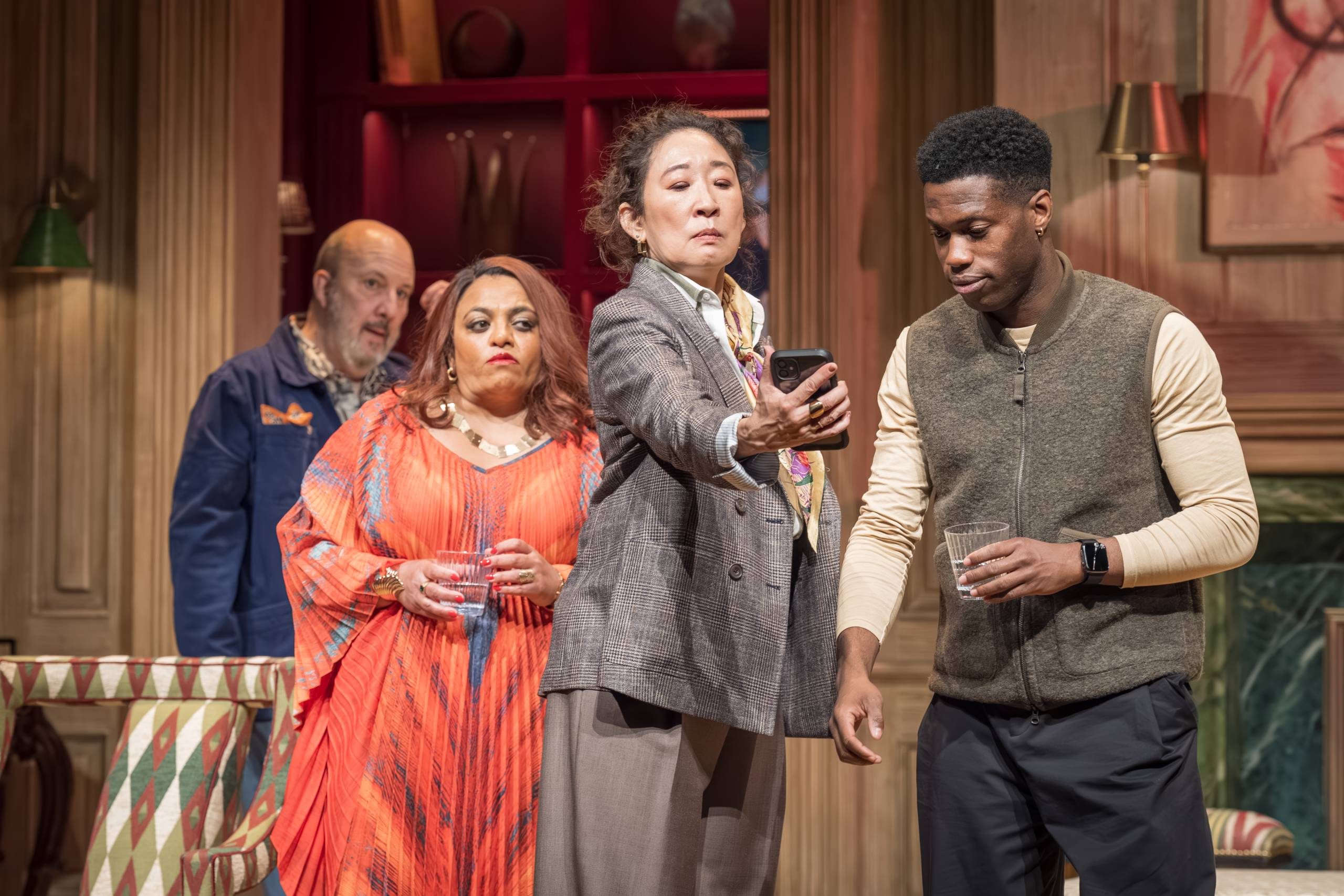 Four people are gathered in a room with elegant décor. The person in the foreground holds a phone, up for two other characters to look at. A fourth person watches from the background.