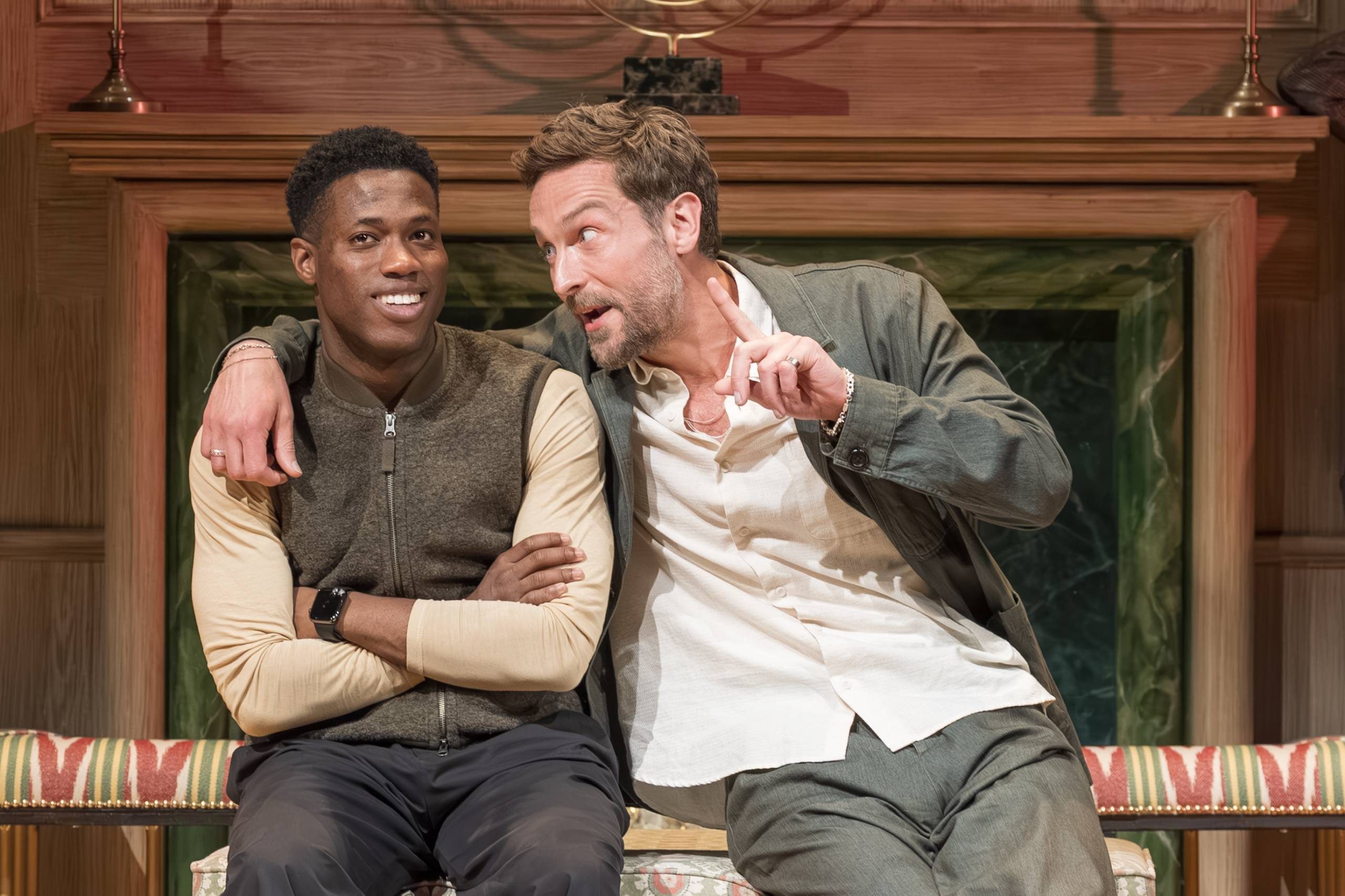 Two people sit together on a colourful patterned sofa in a cosy room. One person looks ahead with a relaxed expression, while the other speaks animatedly, gesturing with a finger. A fireplace with a wooden mantel is in the background.