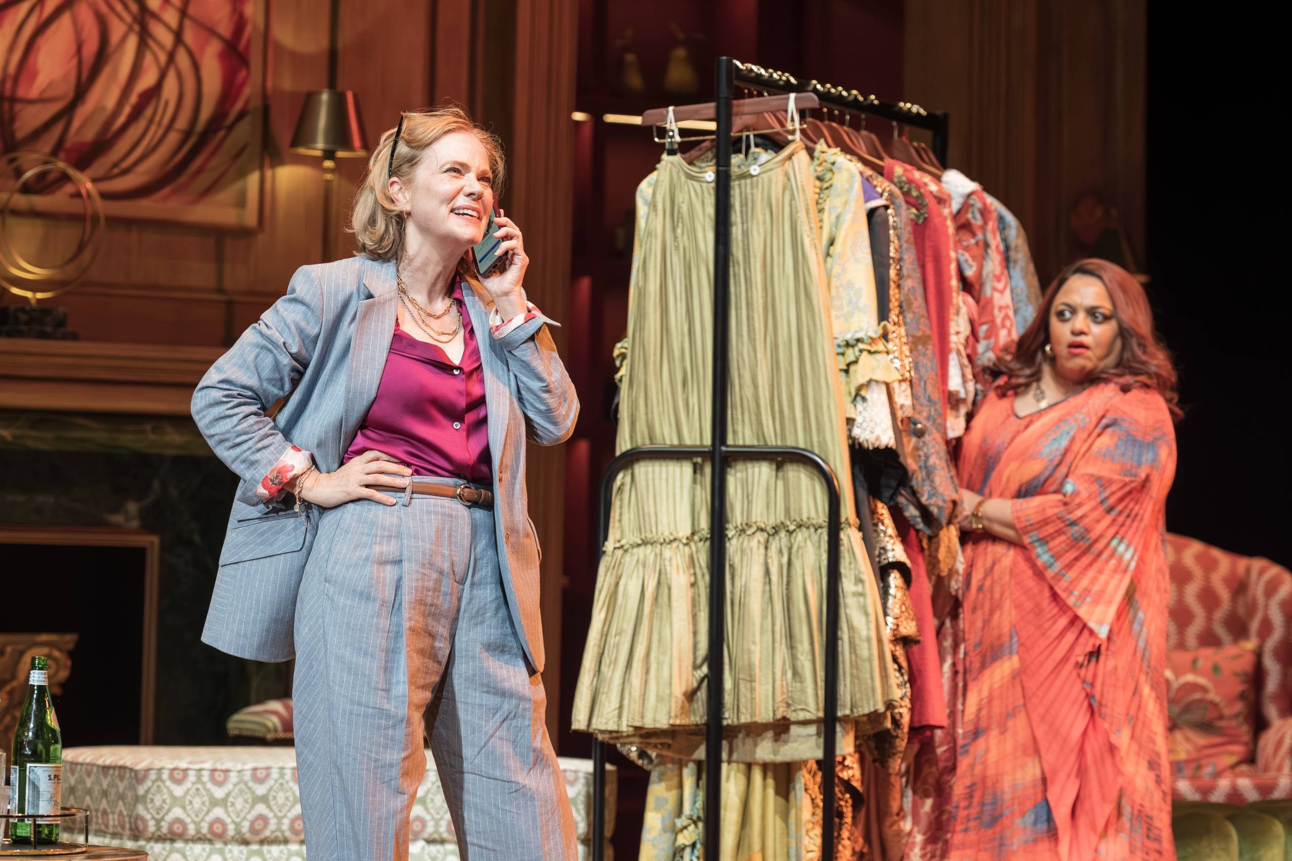 One person is talking on a phone, wearing a light grey suit over a bright top. Another person stands nearby, behind a rack of clothing eavesdropping on the call. The background features elegant décor, including a large abstract painting, patterned furniture, and a bottle on a small table.