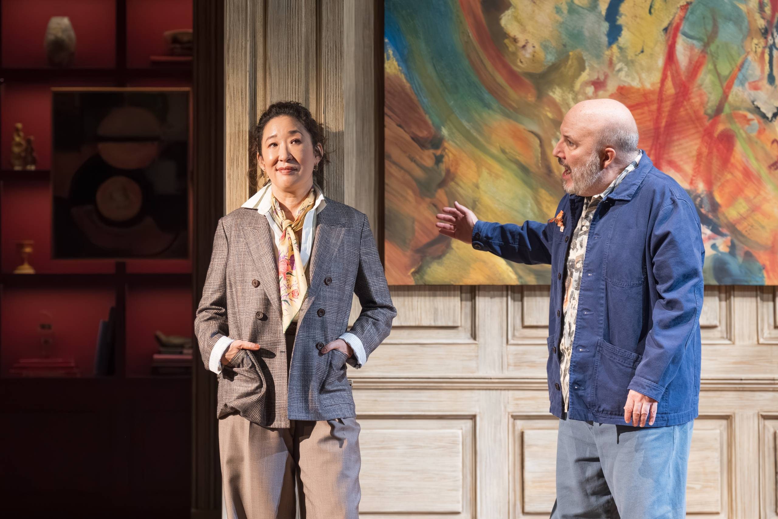One person stands on the left with hands in pockets, wearing a blazer and patterned scarf. The other, on the right, gestures expressively. They are in front of a colourful abstract painting and shelves with decorative items.