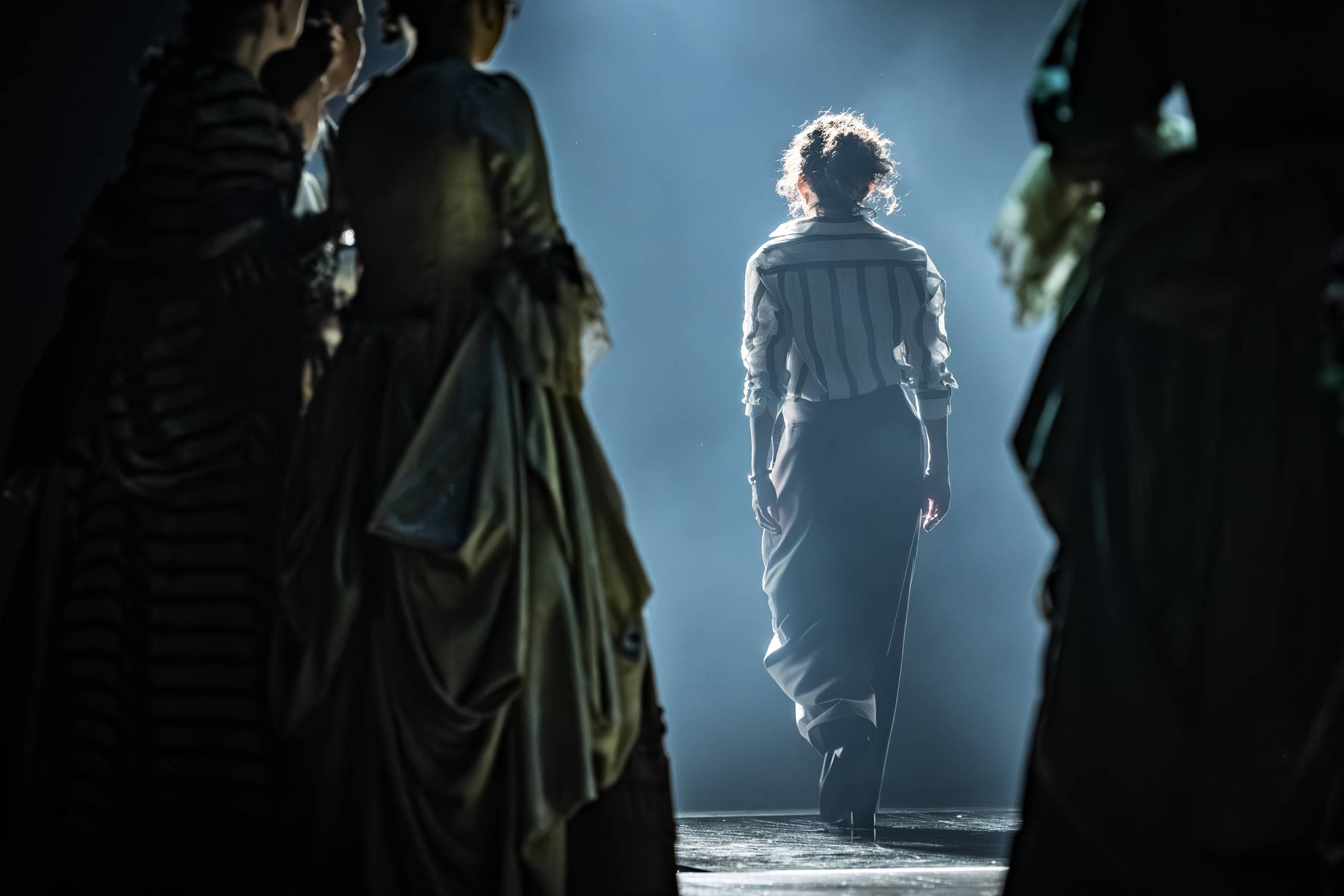 Sandra Oh in a striped shirt and dark pants walks away on a dimly lit stage, surrounded by others in historical attire. Spotlighting creates a dramatic and moody atmosphere.