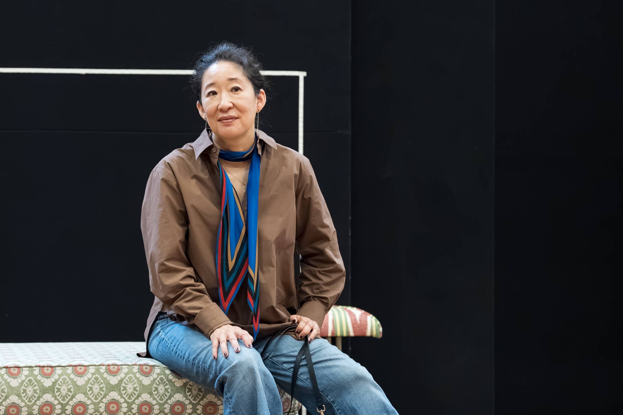 Sandra Oh sitting on a patterned bench, wearing a brown jacket and a colourful scarf, holding a bag.