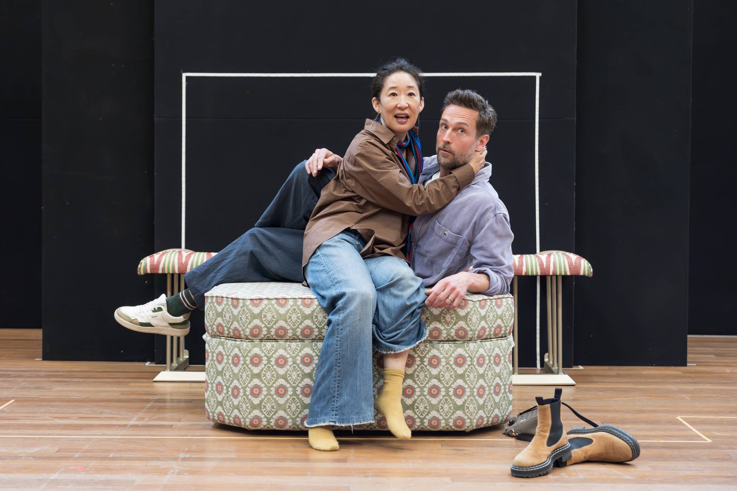 Two individuals are playfully posing on an upholstered ottoman on a wooden stage. One is sitting facing the camera with an arm around the other's neck, while the other person appears relaxed. A pair of brown boots is placed on the floor nearby. The backdrop is a simple black and white design.