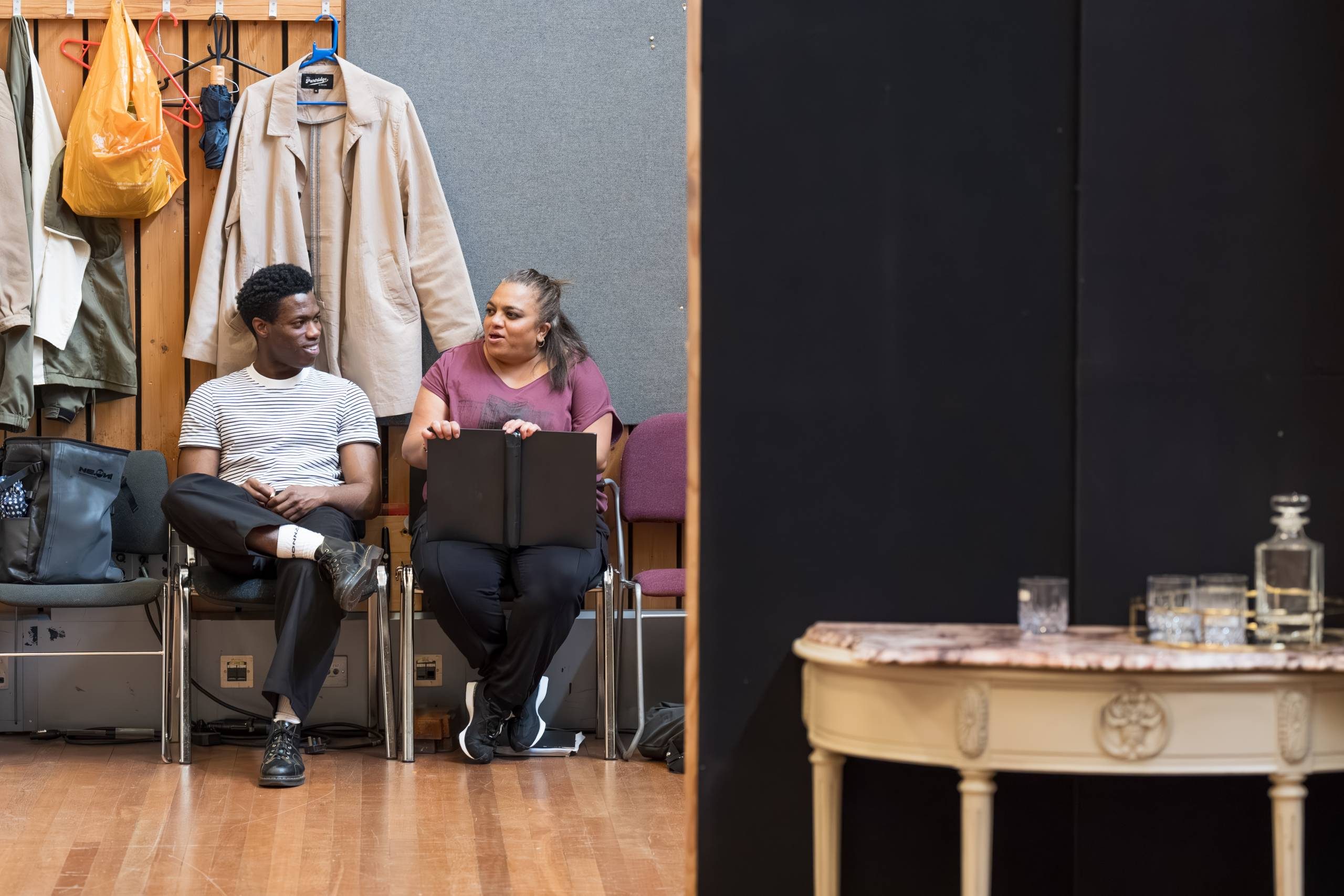 Two actors are seated on chairs in a backstage area, conversing. They are surrounded by coats and bags hanging on the wall. A small table with glassware and a decanter is visible on the right.