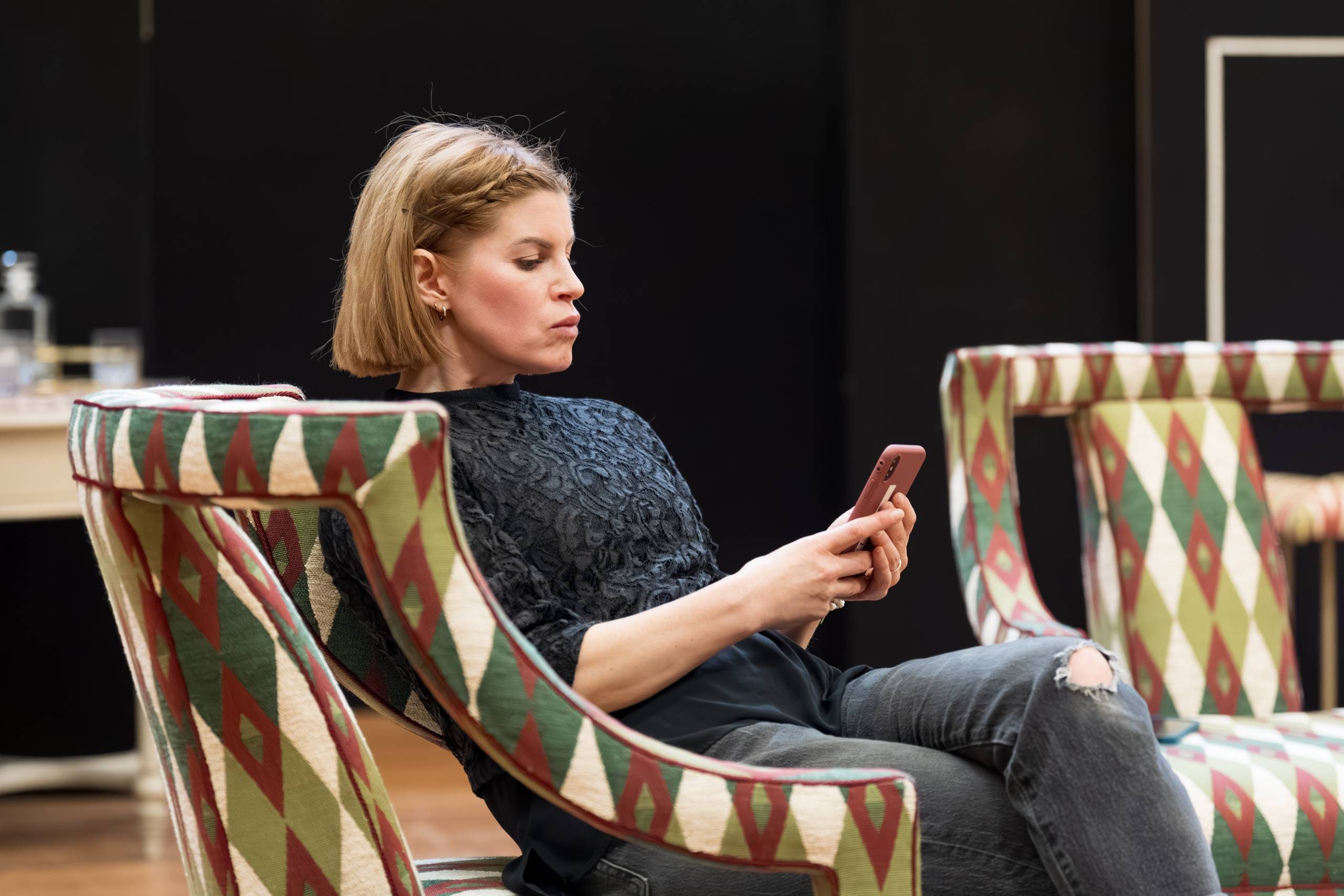 An actor is seated and looking at their phone, surrounded by colourful, patterned chairs
