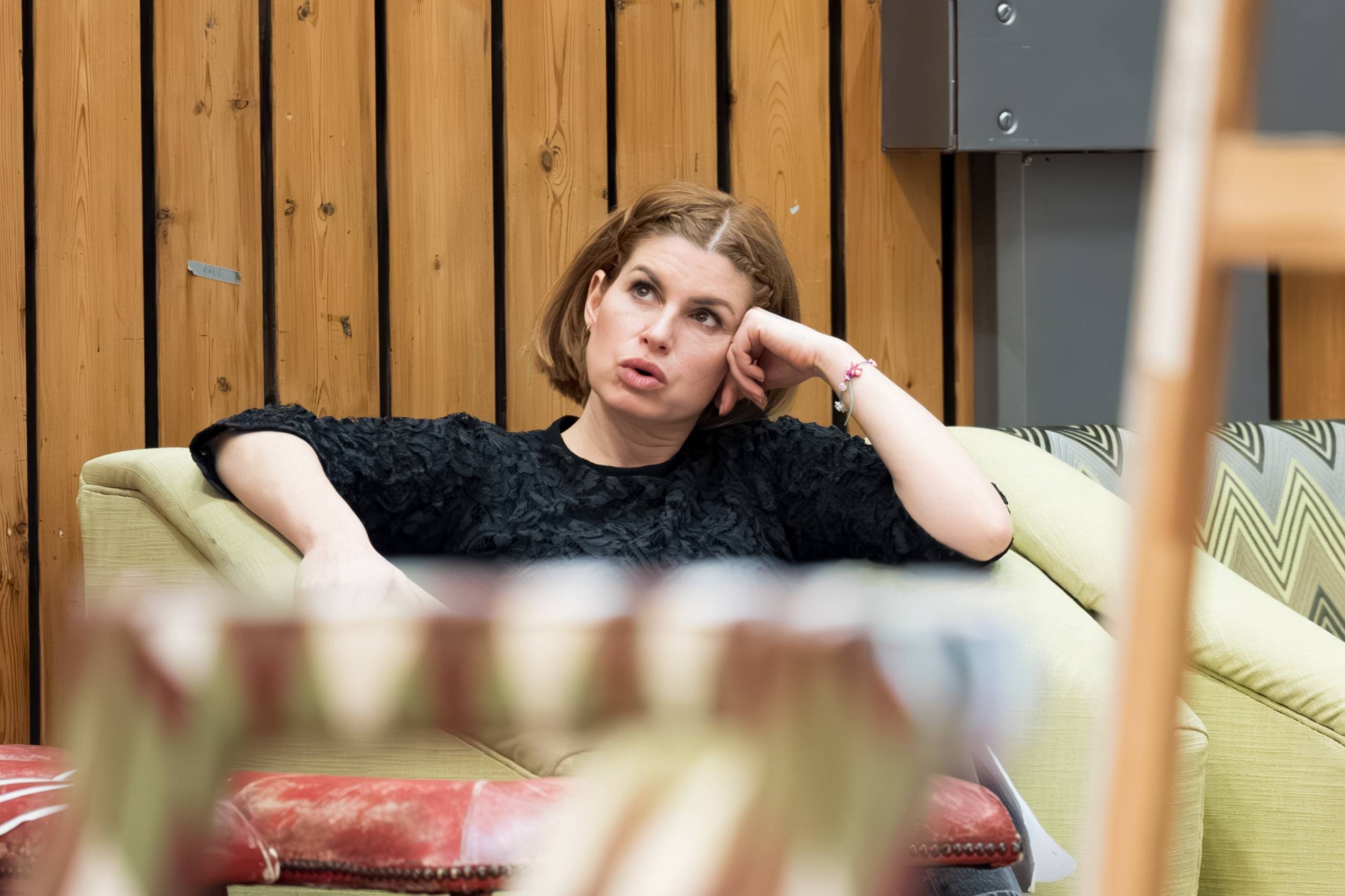 A person sits on a sofa, leaning back with their head resting on one hand, appearing thoughtful. The background features vertical wooden panels and part of a colourful patterned chair is visible in the foreground.