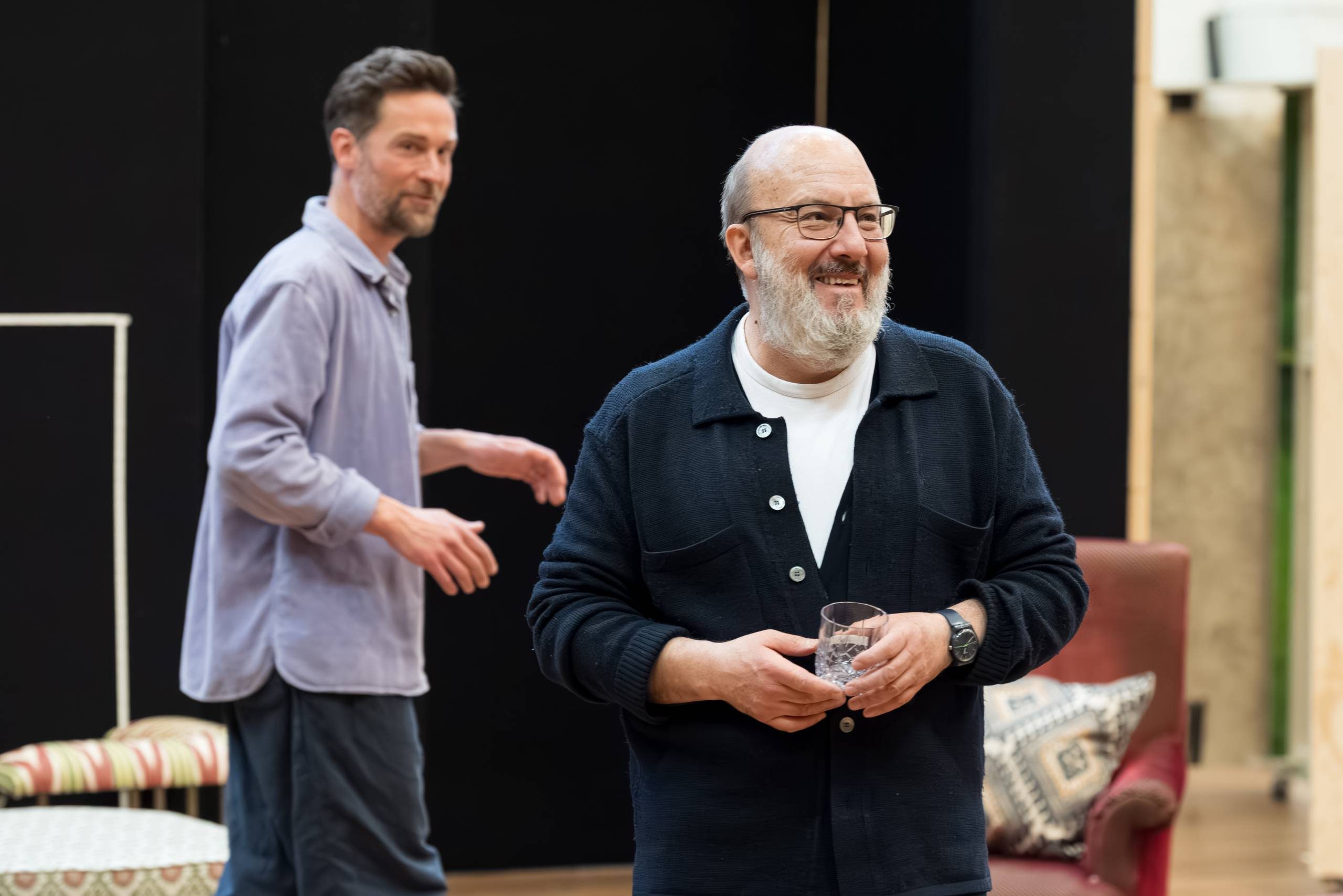Two actors stand casually in a rehearsal space. The actor is the foreground is wearing a half-buttoned shirt and holding a glass in his hands.