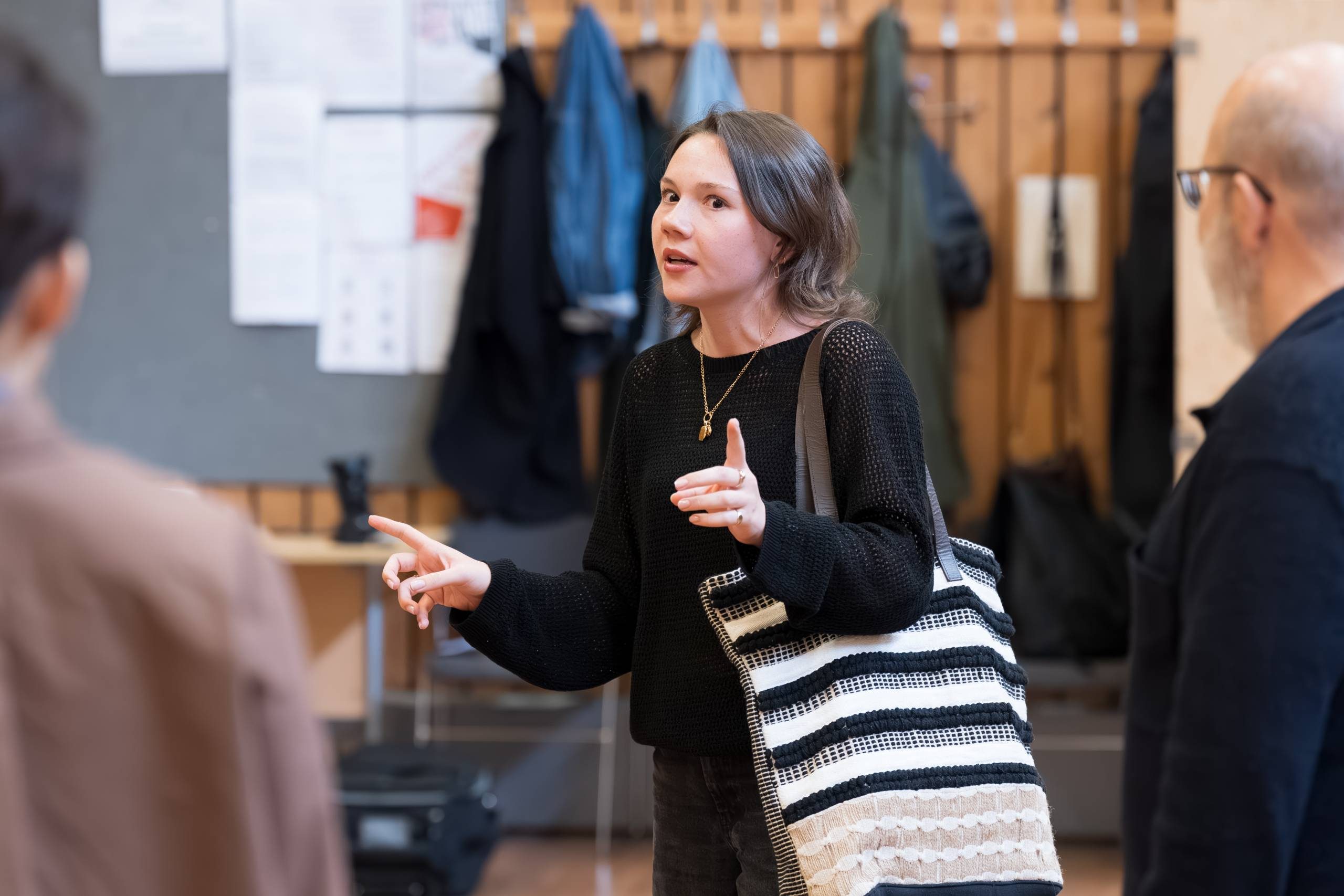 An actor engaged in conversation, gesturing with their hands. They are holding a black and white striped tote bag. Coats are hung on a rack in the background.