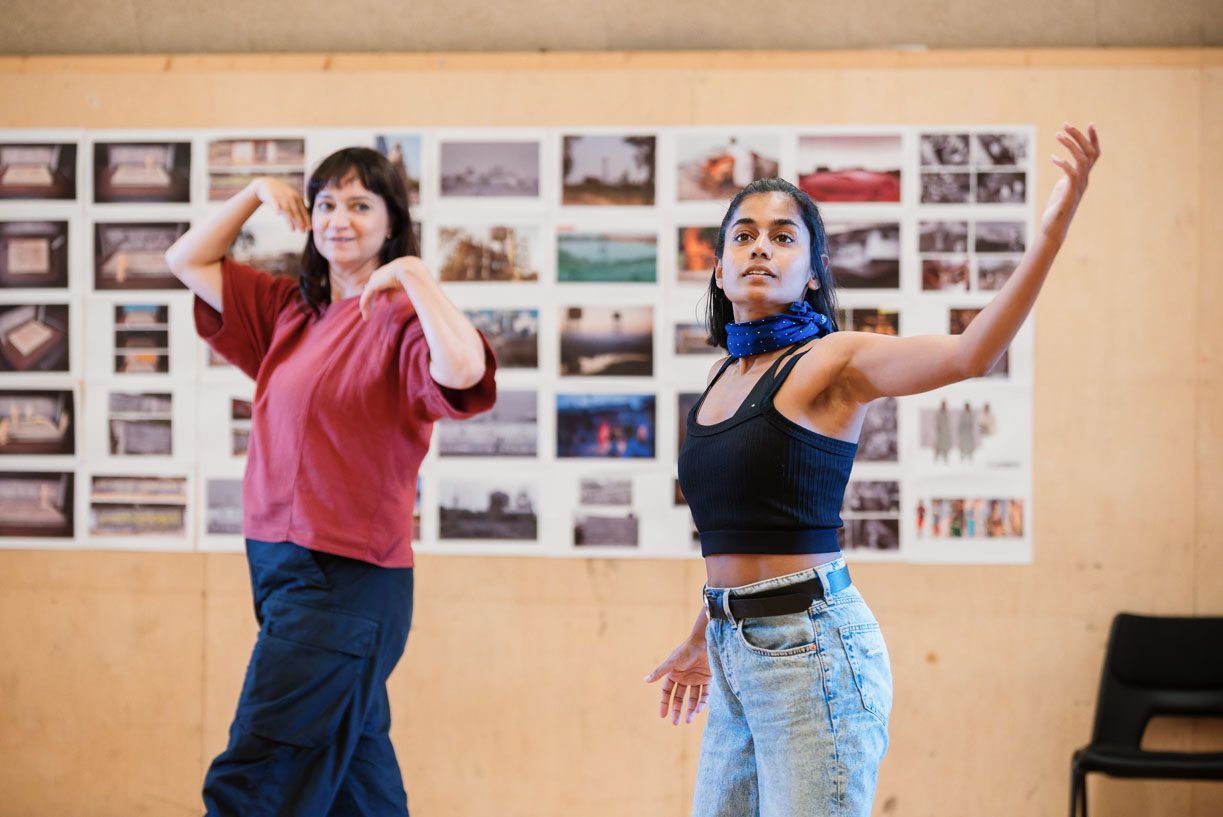 Movement Director Anjali Mehra and Linnea Berthelsen are dancing in a rehearsal room with a wall covered in various photographs. Anjali is wearing a red top and dark pants, and Linnea is in a black top and blue jeans with a blue scarf.