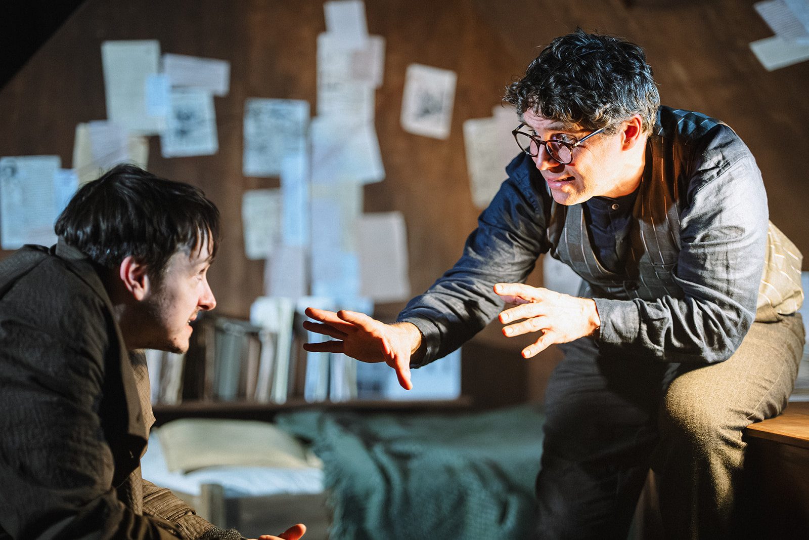 Two men are in a room, they look at each other, one sits on a table slightly above the other, he is holding his hands out toward the other man, as if explaining something.