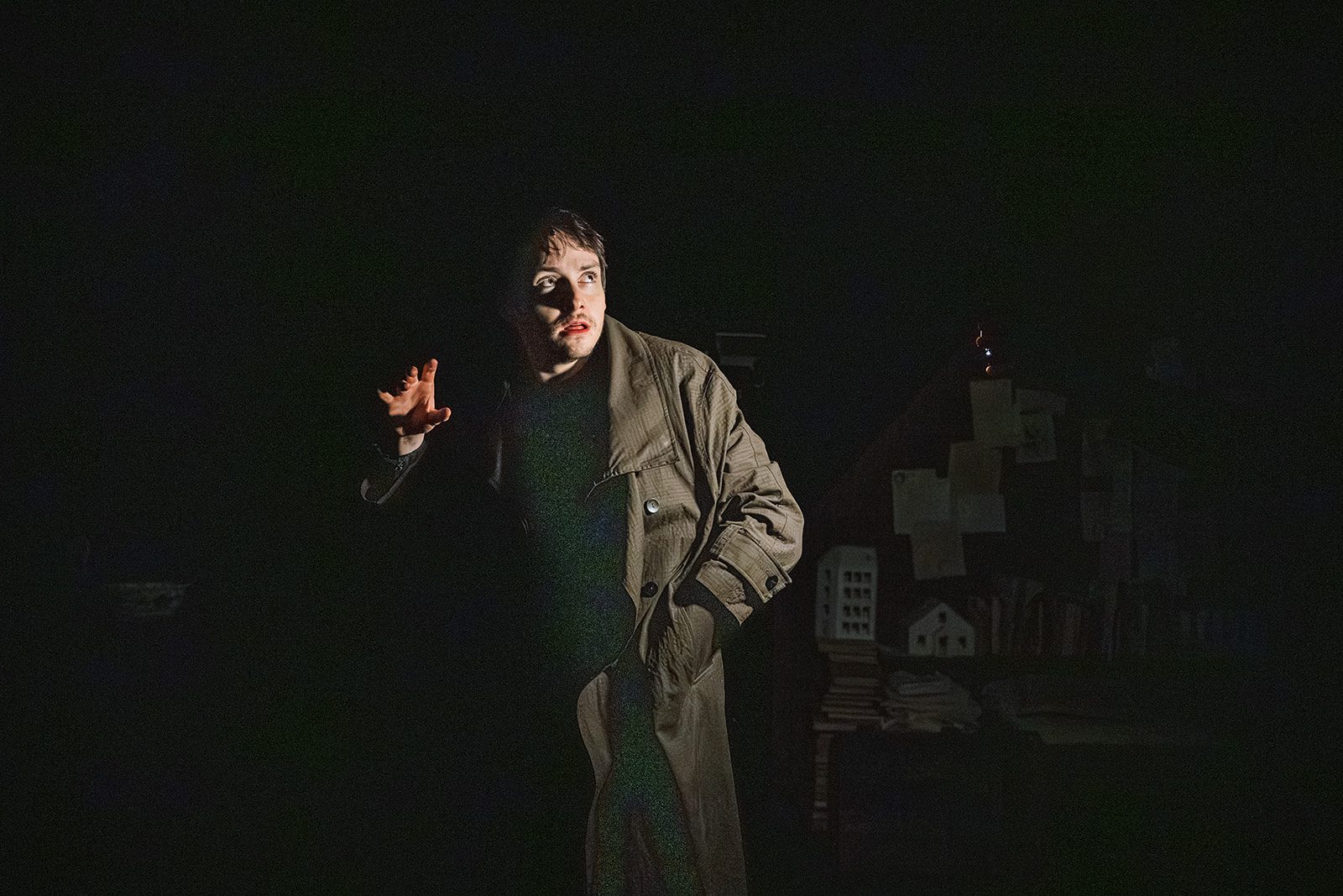 A man wearing a long coat stands in the dark,, he is lit by a light we cannot see, he is the only thing we see. One hand is in his pocket, the other is held up, palm spread wide. He looks frightened and surprised.