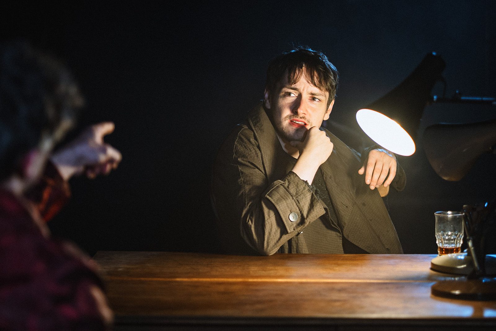 Aman wearing a long coat sits at a table, his elbow rests on the table and his chin rests against his hand. A man sit opposite him with his back to us, he is pointing at the man we can see. The room is very dark, the table and the man we can see are lit by a single lamp.
