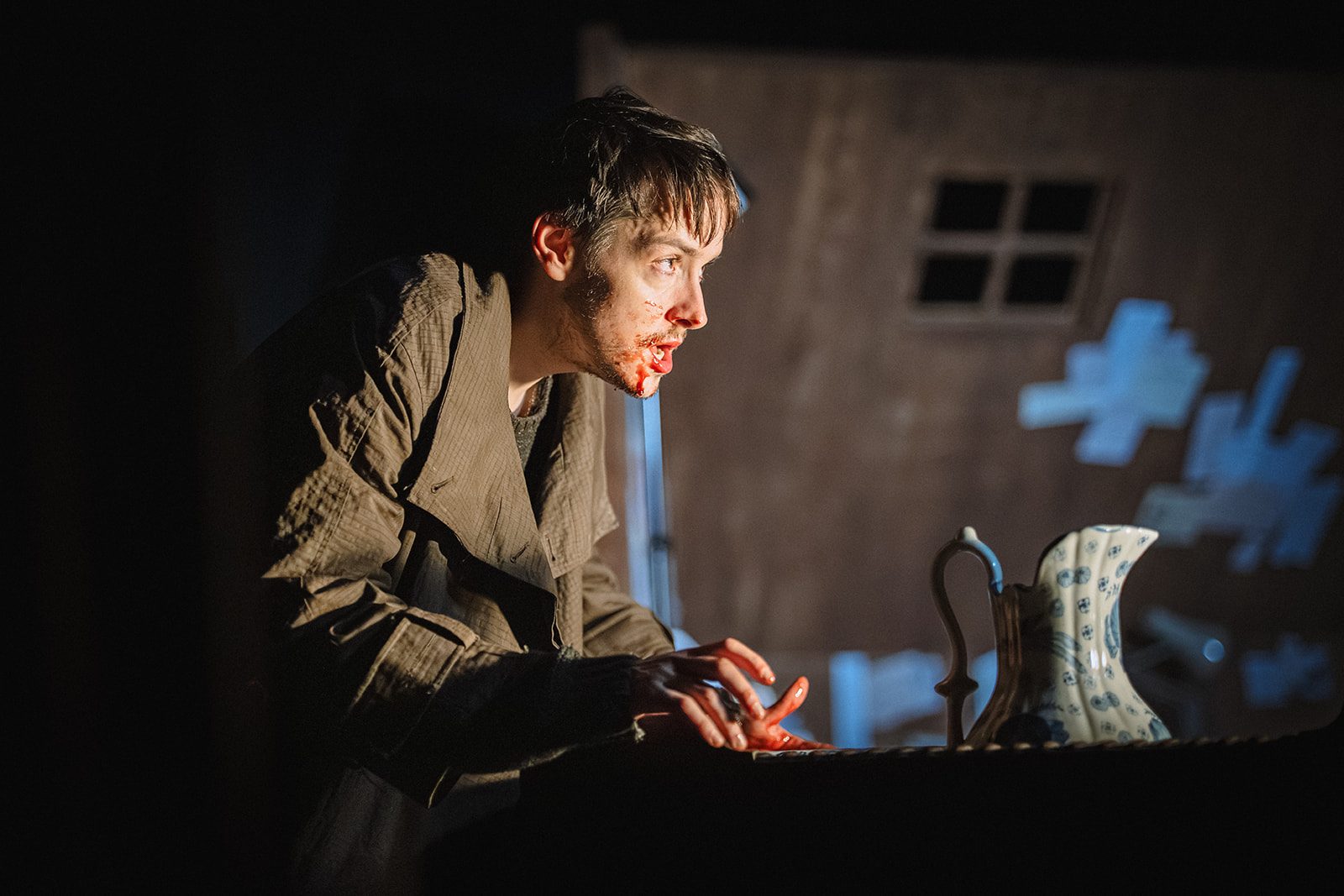 A man is hunched over a table, his face and hands are splattered with blood. A decorated blue and white jug is on the table before him.