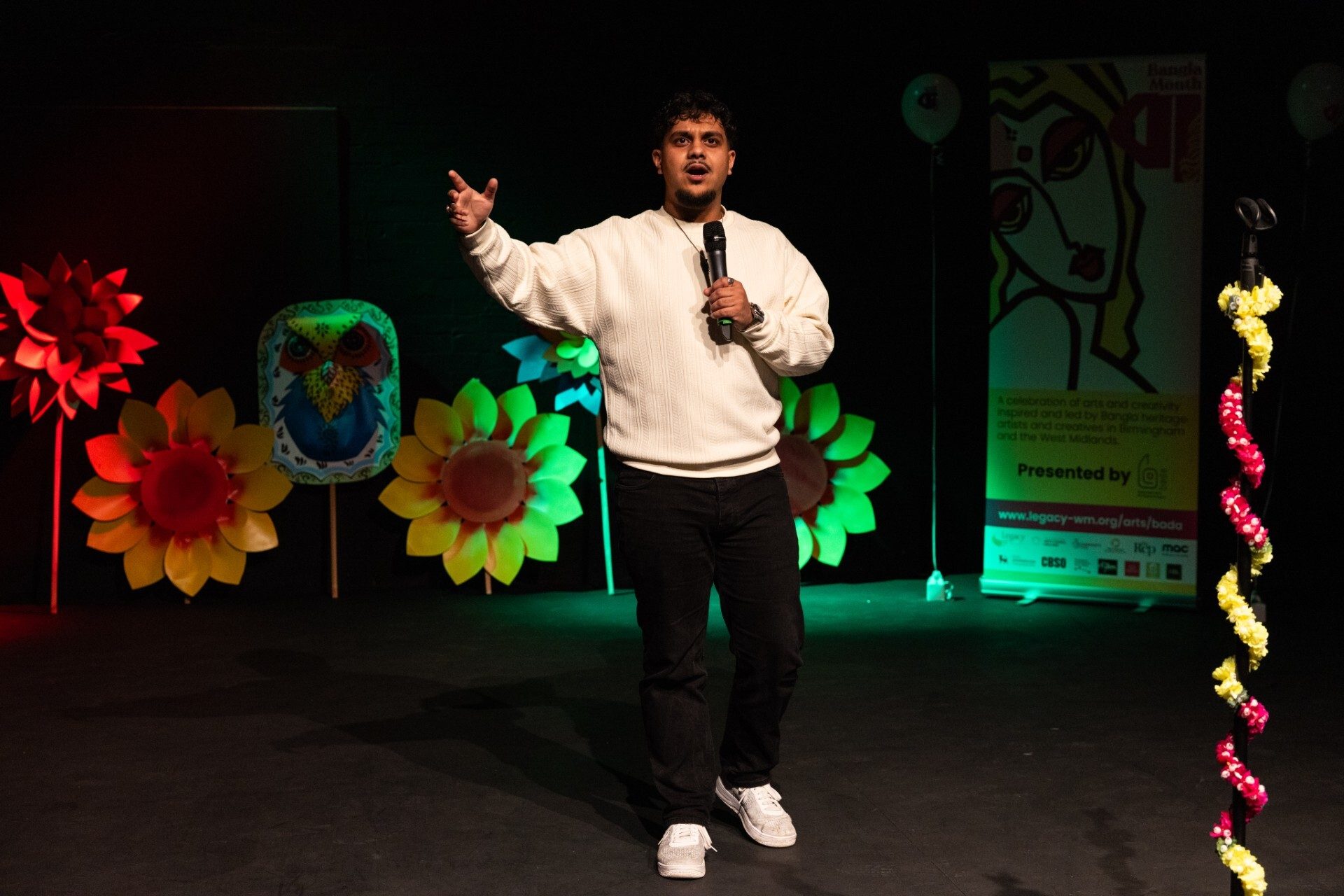 Man in white sweater speaking into a microphone on stage with colourful flower decorations and a banner in the background.