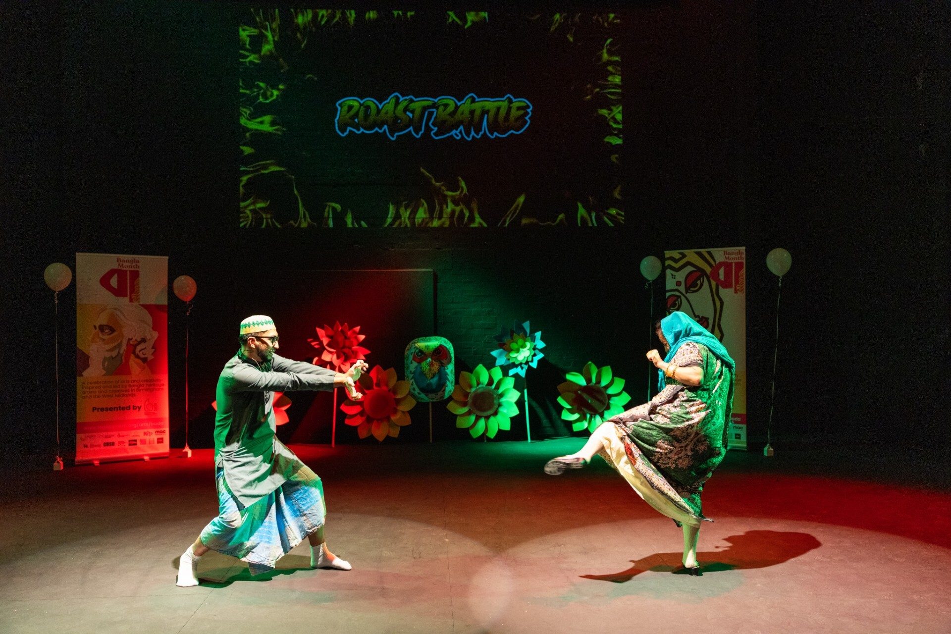 Two performers in traditional clothing face each other on stage with a 