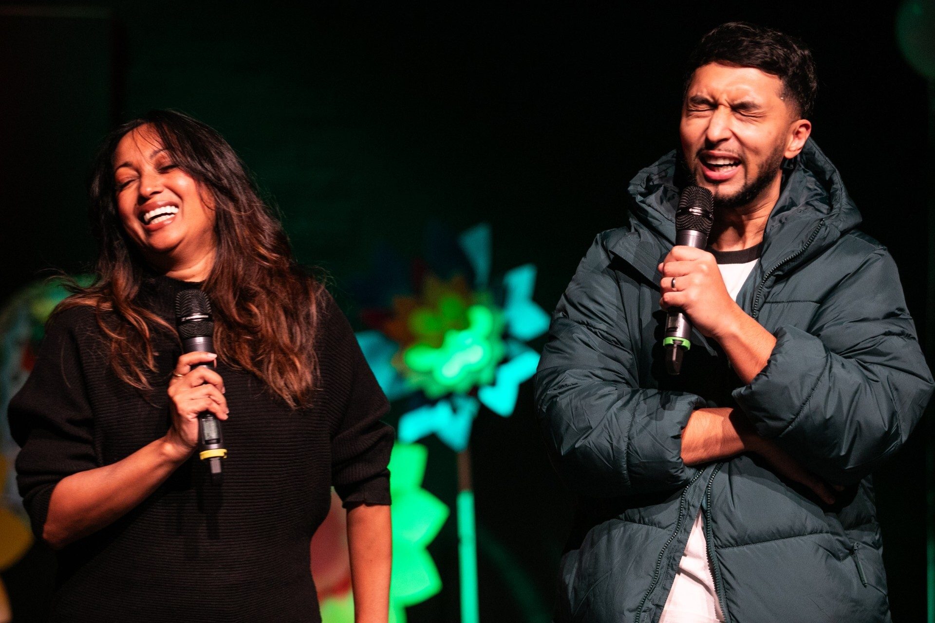 Two people on stage holding microphones, laughing with colourful abstract shapes in the background.