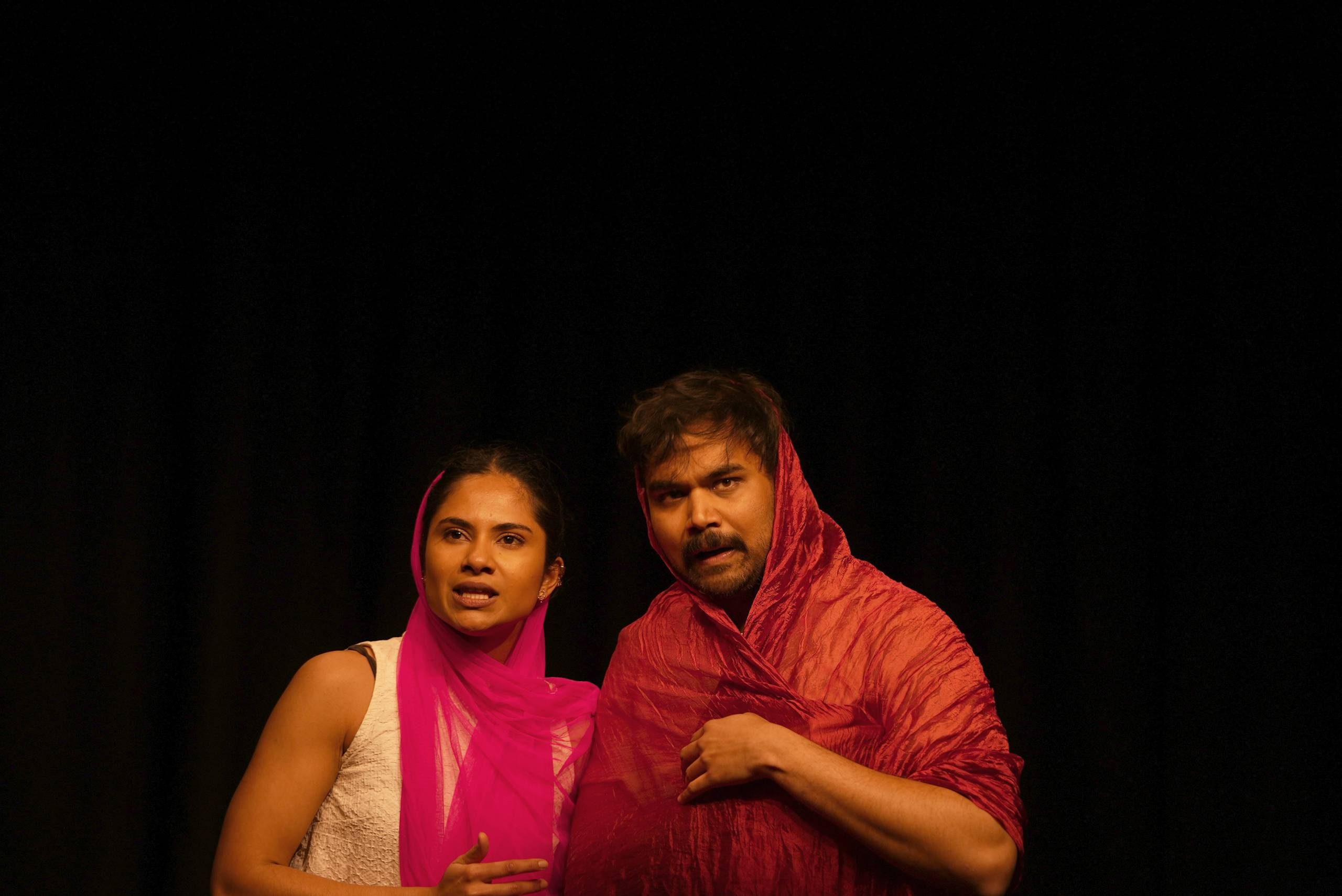 A man and a woman stand against a black background. They are bother wearing headscarves. They are listening to something but seem suspicious about what they are hearing.