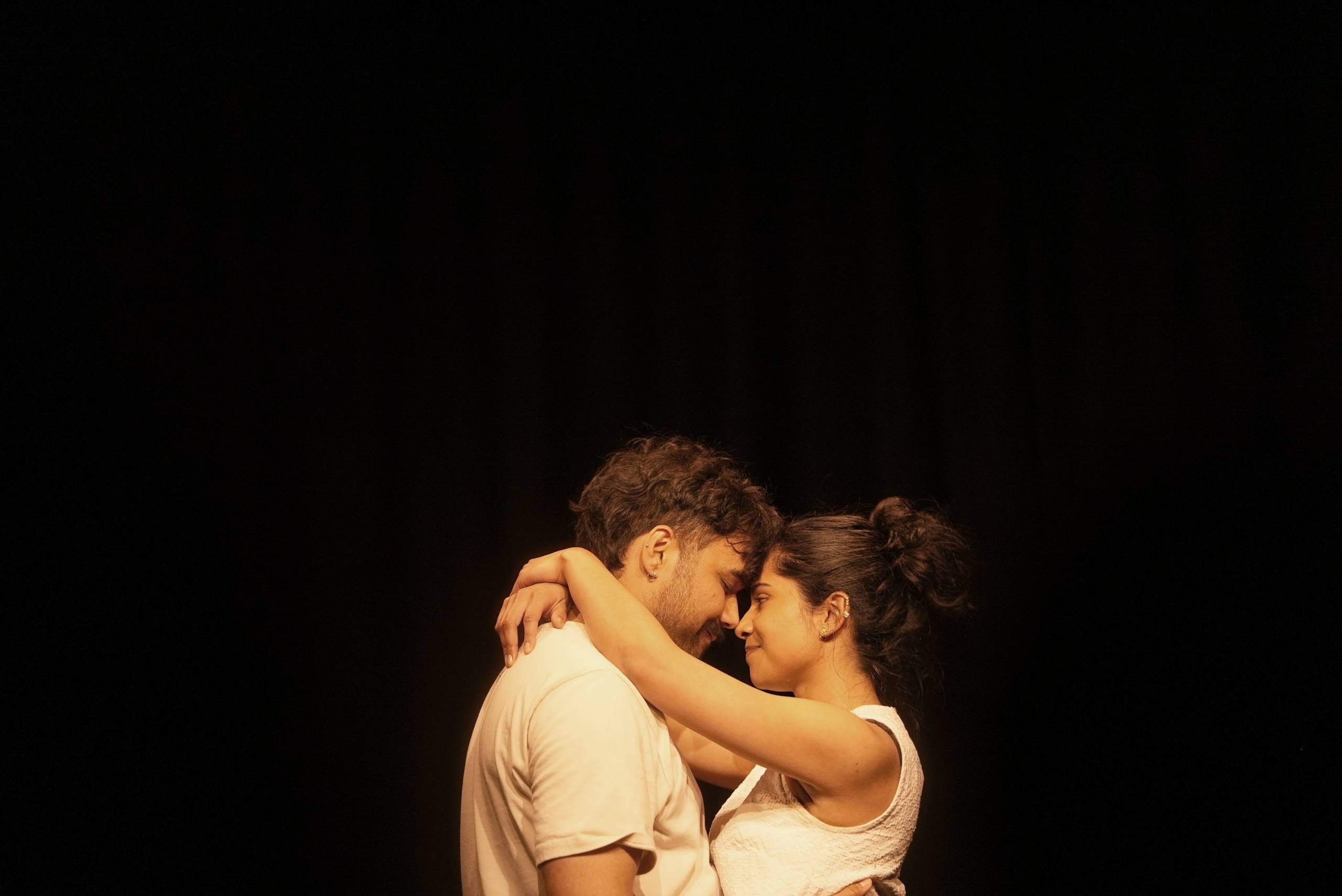 A man and woman stand facing each other against a black background. their noses are touching, her hands are wrapped around his neck, his are wrapped around her waist. They are sharing a moment of intimacy, they both look happy.