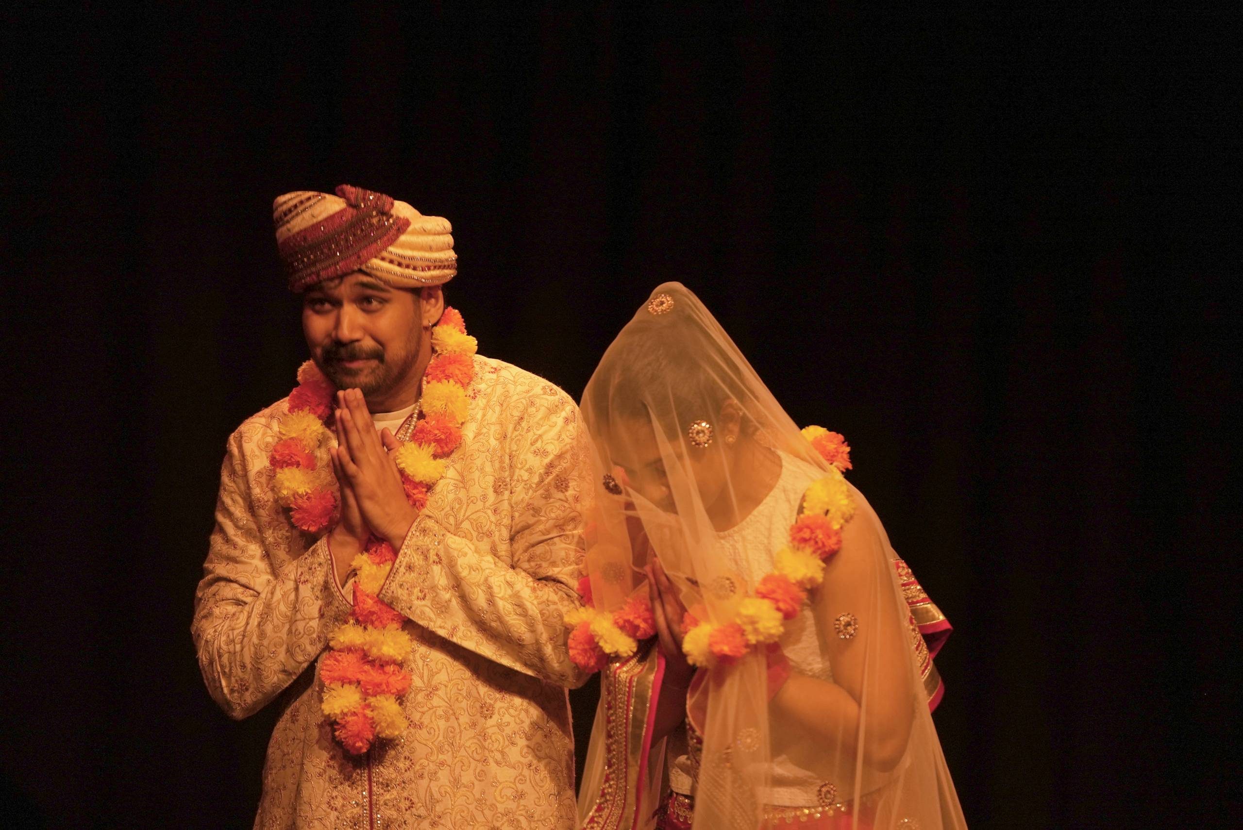 A man and a woman are dressed in Indian wedding dress. He wears a garland around his neck, and his hands are clasped together in prayer, he is smiling. The woman's head is bowed, her face is covered by a veil, her hands are also clasped in prayer.
