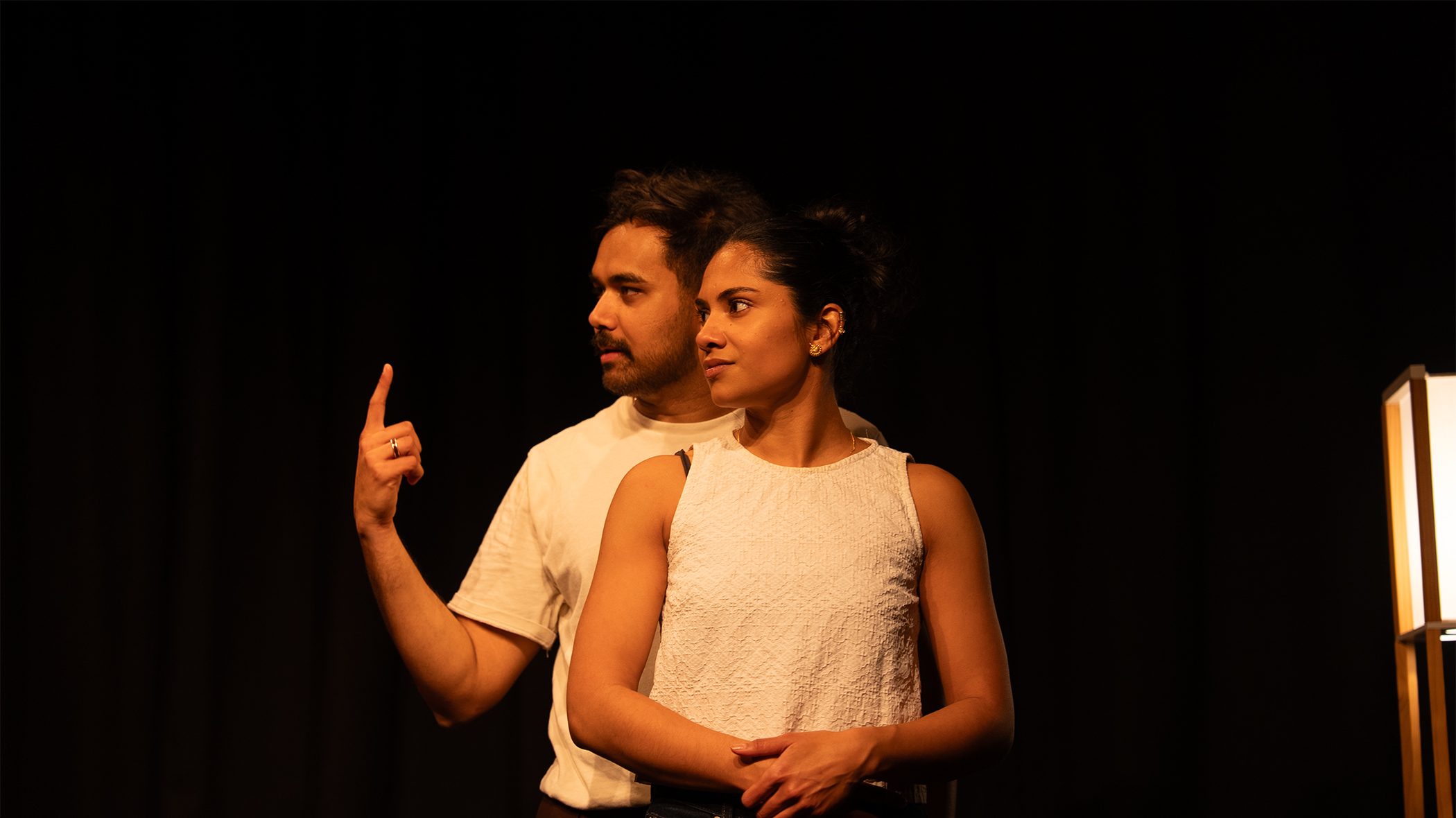 A man and a woman stand together, the man stands behind the woman, one arm wrapped around her waist, the other is raise and he points a finger upward. The woman clasps the mans hand at her waist. They are both looking off to the left of the image, away from the camera.