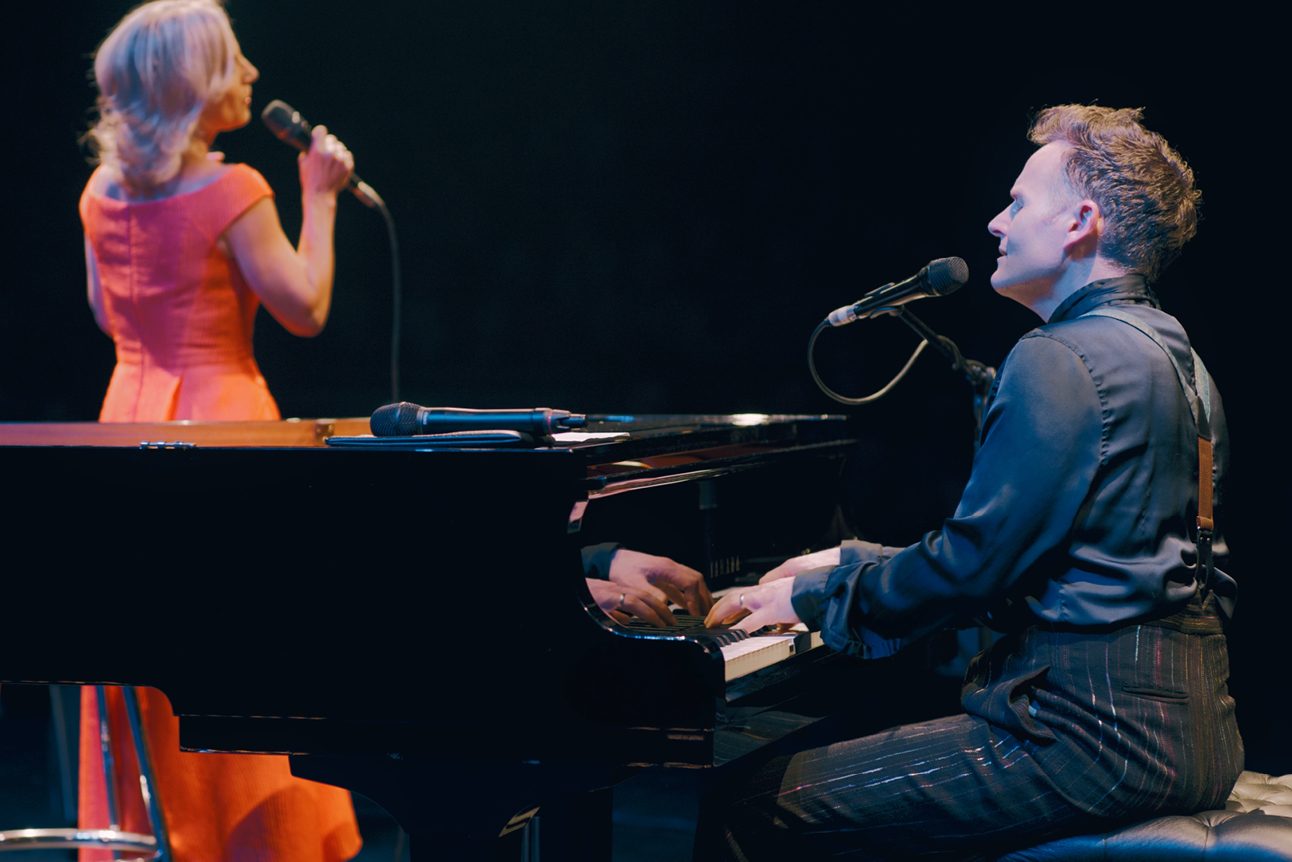 A man in a black outfit is sitting playing a piano and singing into a microphone attached to the piano. A woman in an orange dress is standing, singing into a microphone that she is holding They have their backs to the camera.