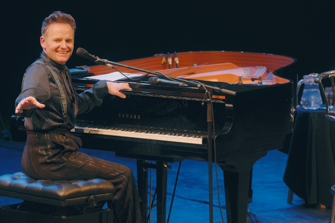 Aman dress all in black is sitting in front of a piano, he is turning toward the camera and smiling.