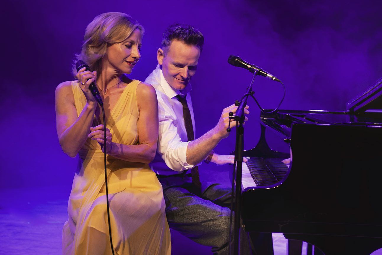 A man wearing a white shirt, black tie and trousers and a woman wearing a yellow dress are sitting together in front of a piano. The man is playing the piano, the woman is holding a microphone