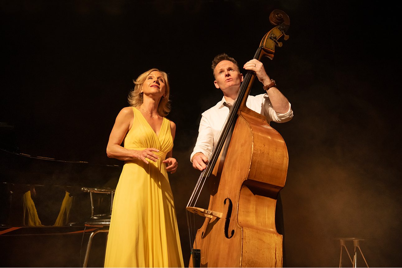 A man wearing a white shirt is playing a double bass, a woman wearing a yellow dress stands next to him. They are both looking upward.