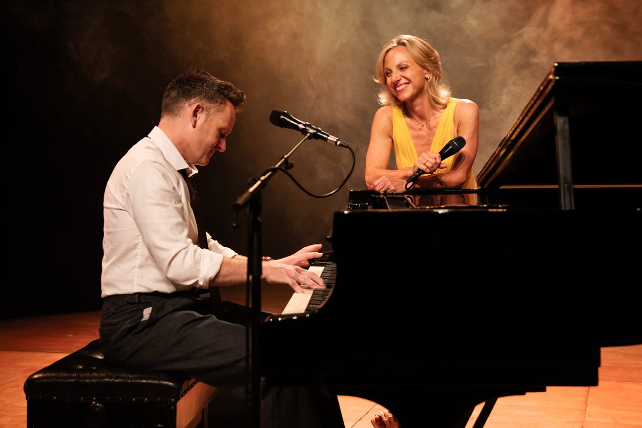 A man wearing a white shirt, black tie and black trousers is playing a piano. A woman wearing a yellow dress is smiling and leaning against the piano looking at the man, she is holding a microphone