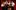 The Clowns stand together in a line in front of a curtained backdrop, they hold microphones and each has a different expression on their face.